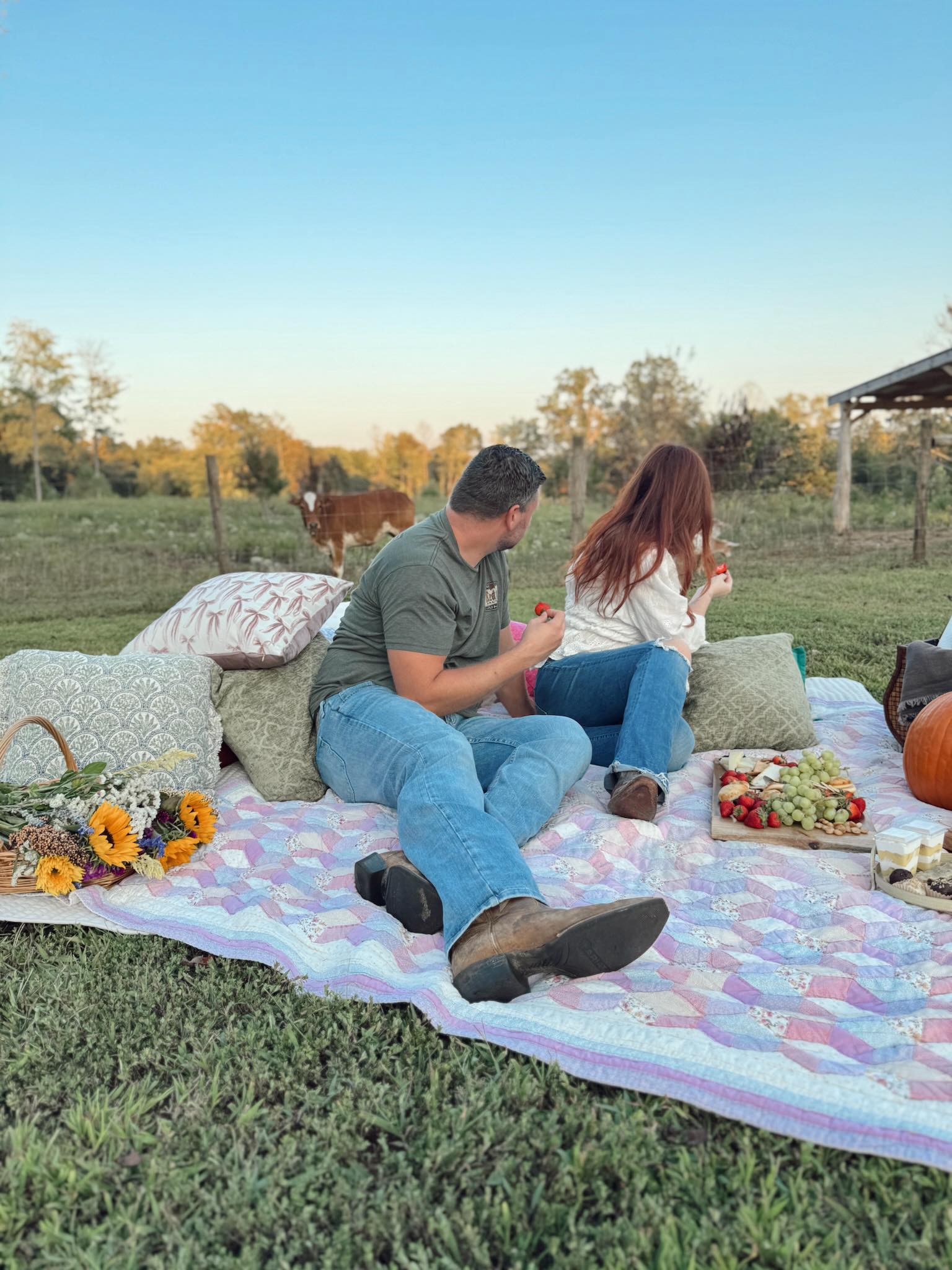 A couple sitting on a quilted picnic blanket at golden hour with sunflowers and a snack spread, a cow grazing in the background