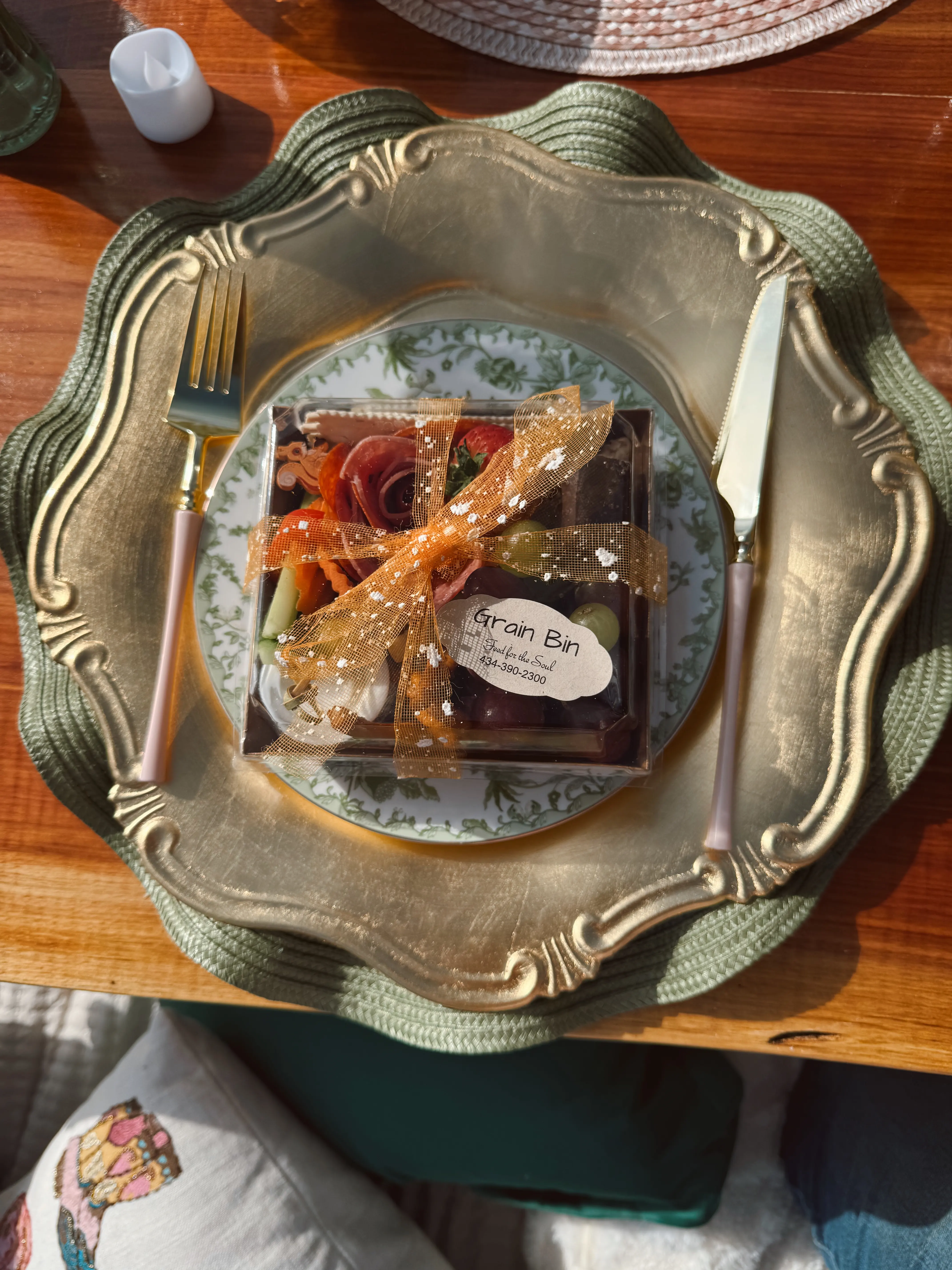 A single-serve Grain Bin charcuterie box tied with a ribbon, presented on a decorative charger plate with gold utensils