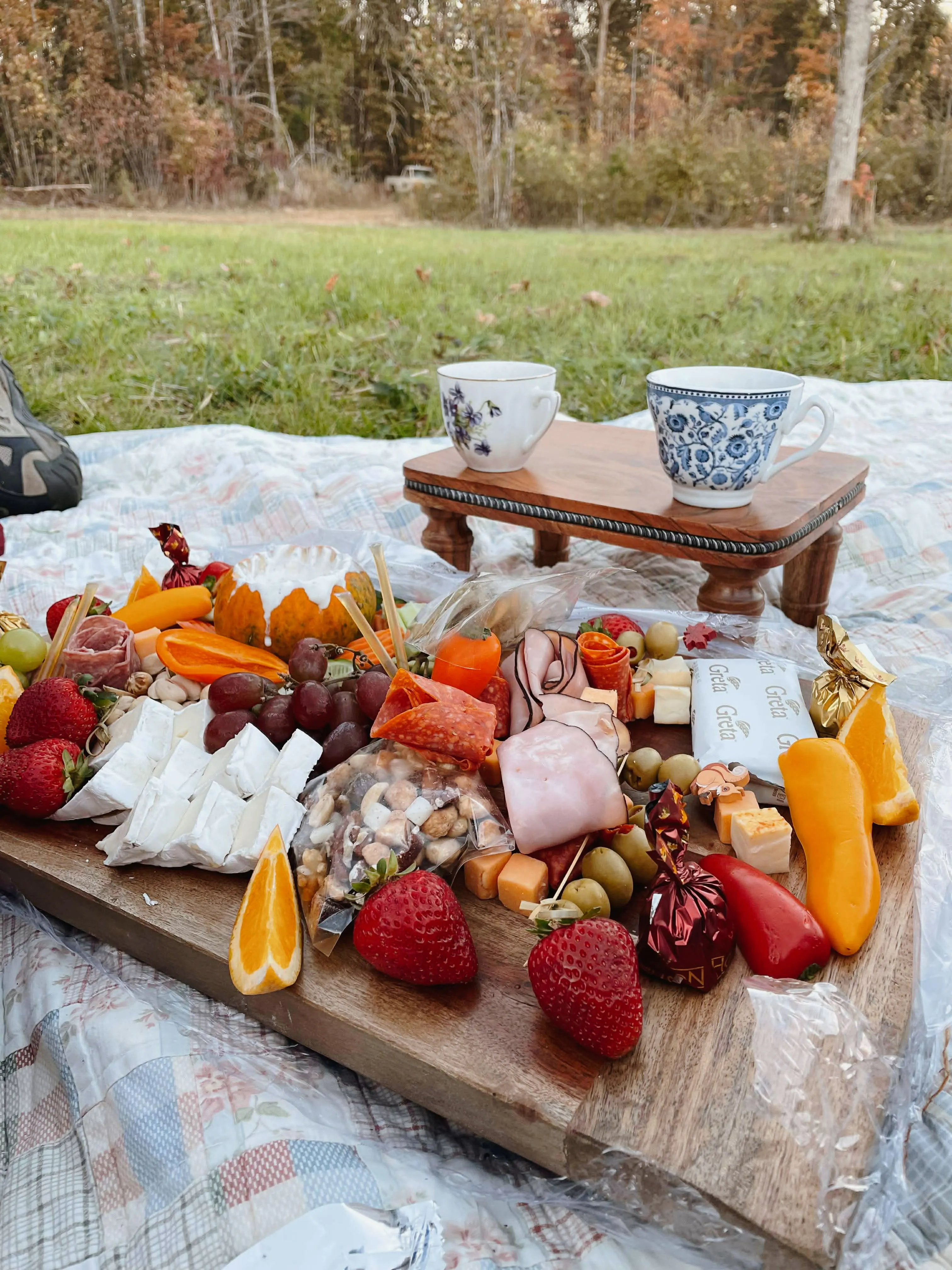 An abundant outdoor charcuterie board with fresh fruits, meats, cheeses, and crackers, with china teacups and fall foliage