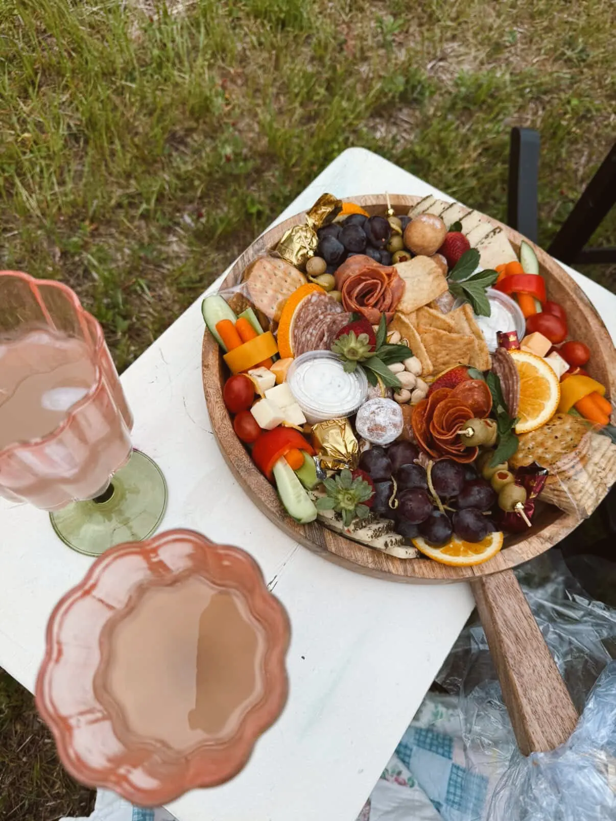 A round wooden charcuterie board with salami roses, grapes, crackers, and dips alongside blush-colored glasses