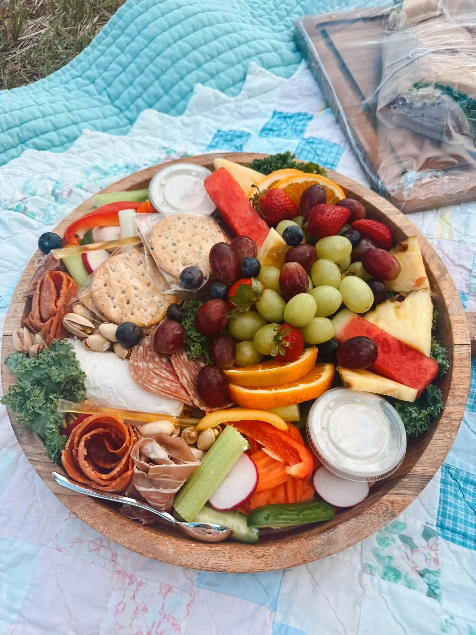 A round wooden charcuterie bowl loaded with fresh fruit, crackers, meats, and dips on a quilt picnic blanket