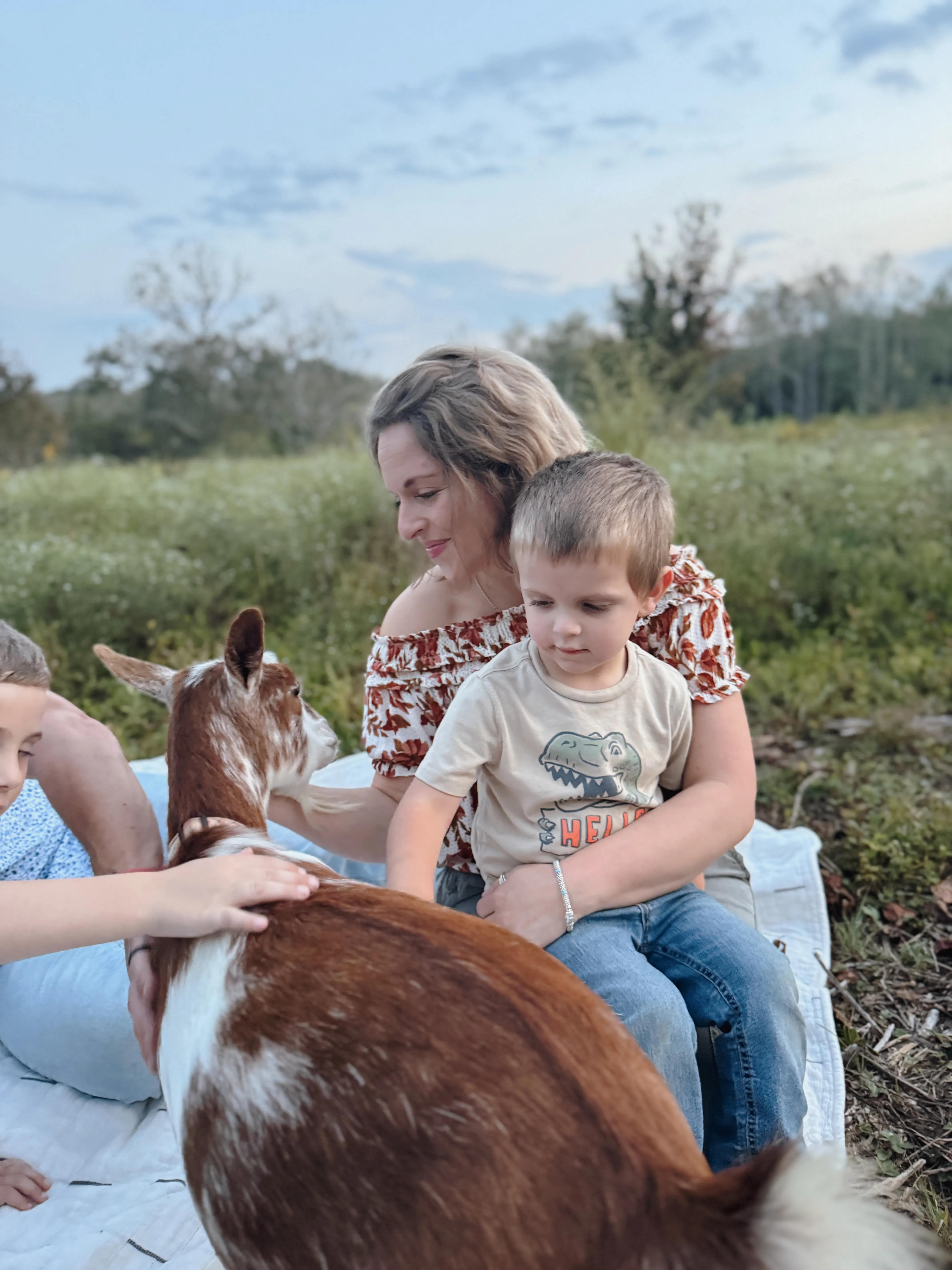 A mother and her young son sitting on a picnic blanket at dusk while baby goats roam nearby at Magnolia Hill