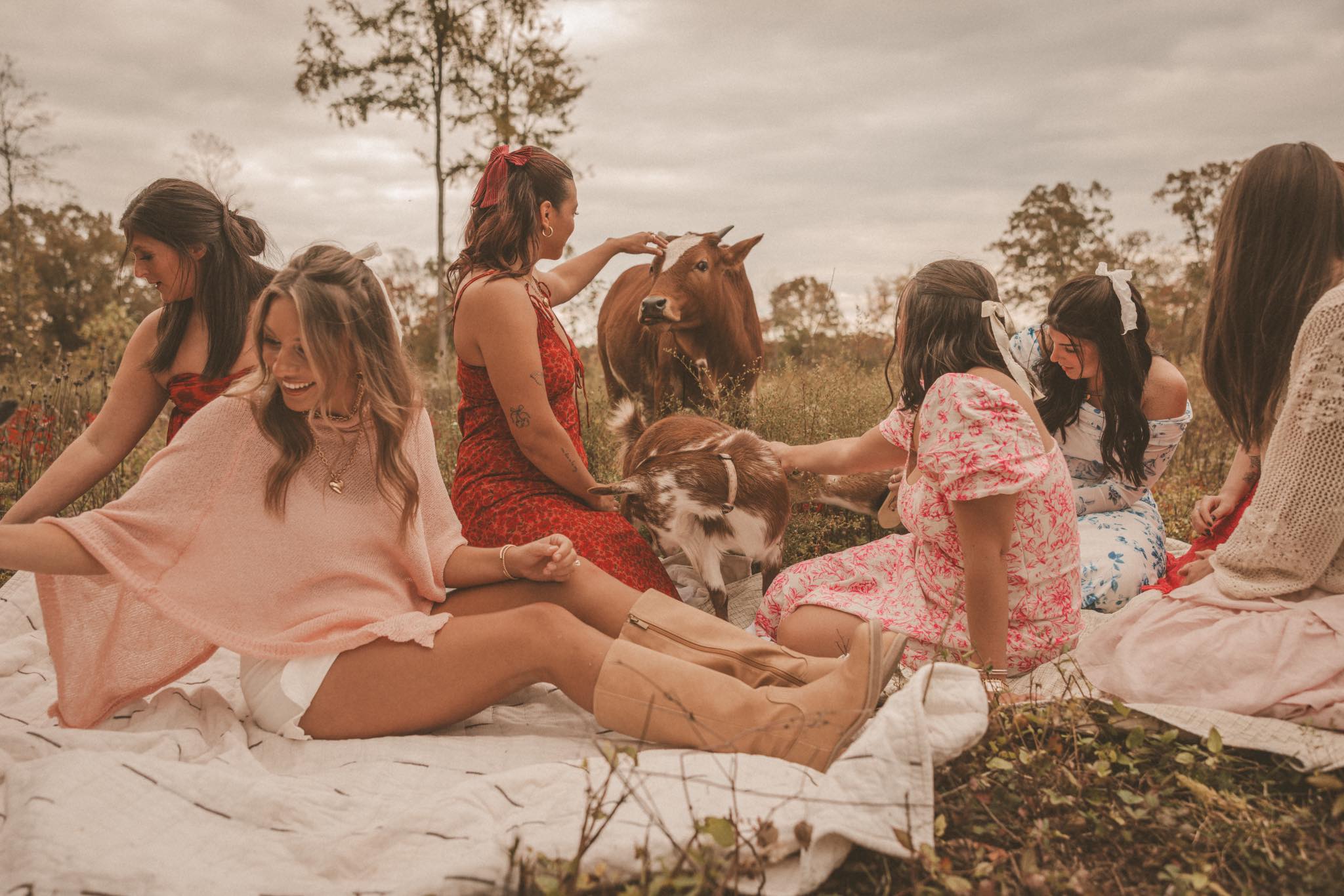 Women in colorful dresses laughing and petting a goat on a picnic blanket in an autumn field at Magnolia Hill